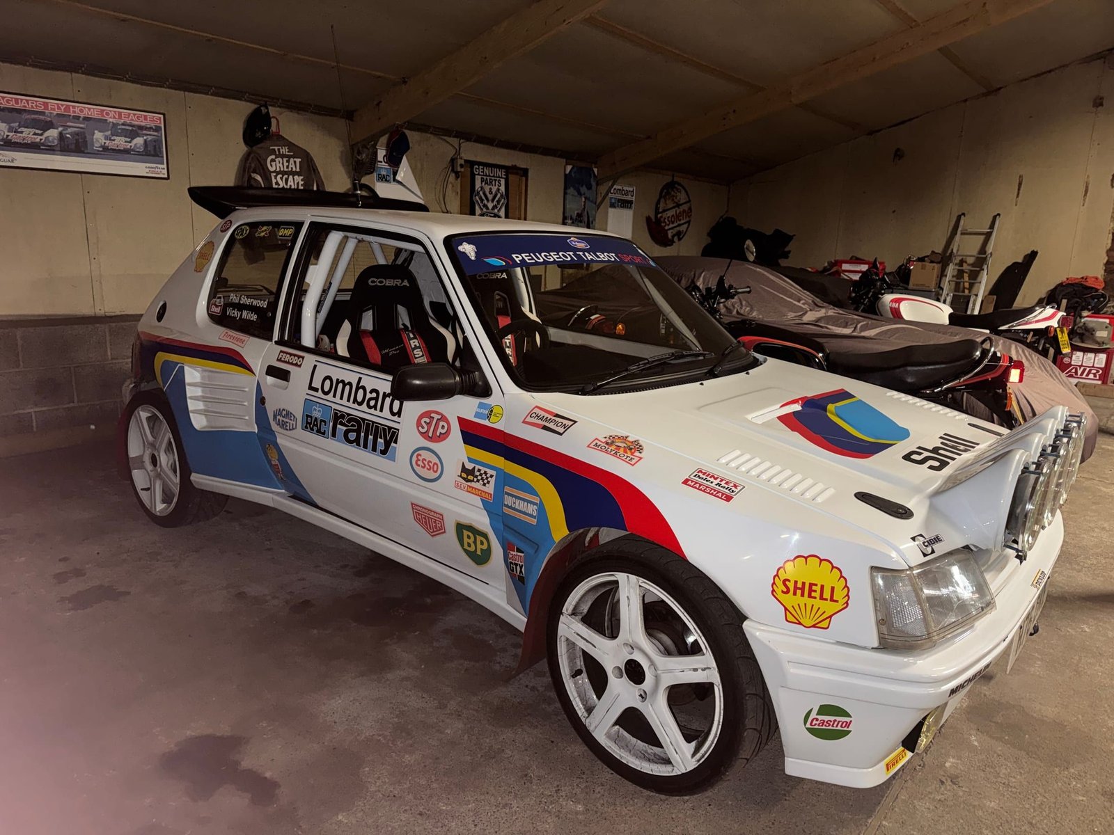 White Peugeot 205 GTI in period Lombard RAC Rally livery with period sponsor decals, parked in a garage.