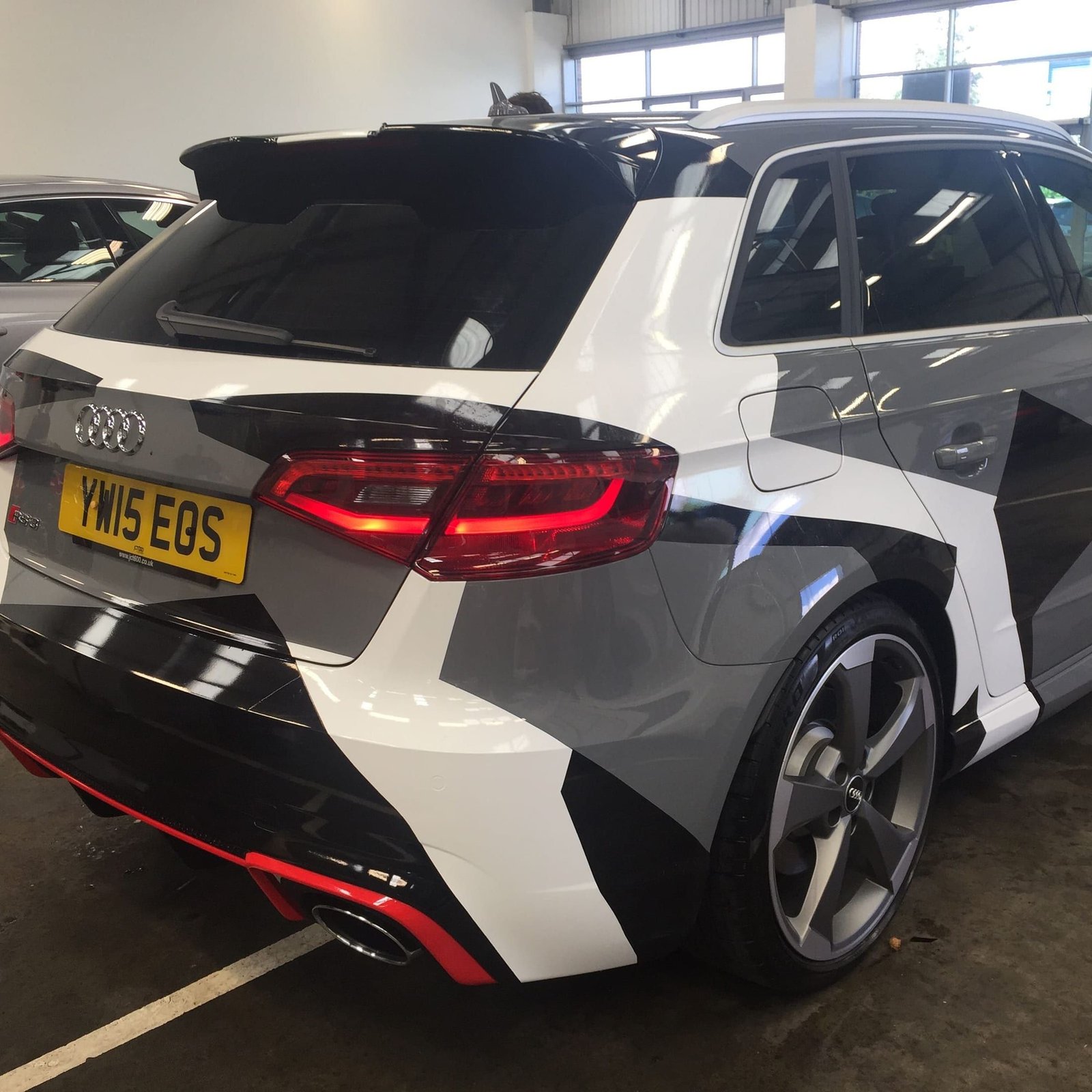 Audi RS3 estate with a grey, white and black geometric camo-style vehicle wrap, rear three-quarter view.